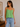 Woman wearing a green lace top and blue jeans in a room with a wooden shelf and framed picture.