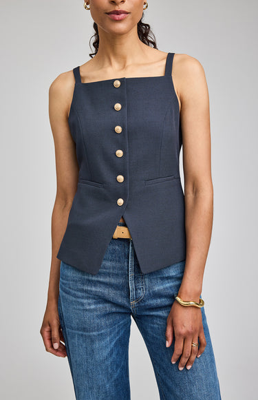 Woman wearing a navy blue sleeveless top with buttons and blue jeans on a gray background
