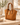 Camel suede handbag with gold hardware shown from a side angle on a neutral travertine table.