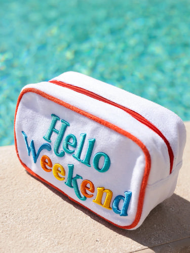 White pouch with 'Hello Weekend' text in colorful letters, placed on a sandy surface with a pool in the background.