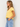A woman wearing a light yellow short-sleeve V-neck crop top and light blue jeans, standing against a plain off-white wall.