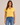 A woman modeling a short-sleeve yellow V-neck crop top paired with blue jeans, set against a plain beige studio background.