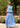 Woman wearing a blue tank and skirt standing outdoors with greenery and flowers in the background