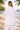 Person in a white outfit standing on a beach with palm trees and a gazebo in the background.