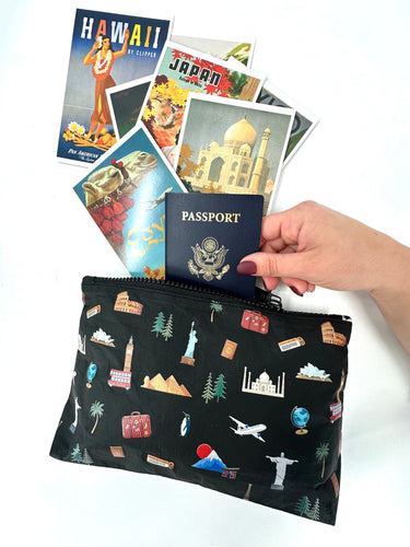 A cosmetic zipper pouch in black with world landmark pictures printed onto it a woman putting a passport and postcards into it