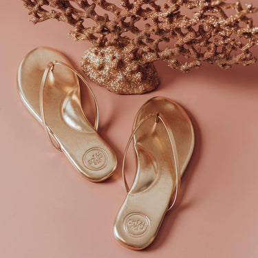 Metallic Gold flip-flops on a pink background with decorative elements.