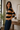 A woman wearing a long-sleeve rugby-style knit top with wide horizontal stripes in tan and black, paired with dark blue jeans, standing in a softly lit indoor room with a window, potted plant, and wooden table in the background.