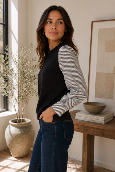 a woman wearing a black crew neck with black and white polo stripe sleeves and blue jeans on neutral background.