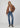 A woman modeling a brown suede-like cropped jacket over a white top, paired with blue jeans and brown heeled ankle boots, against a light gray studio background.