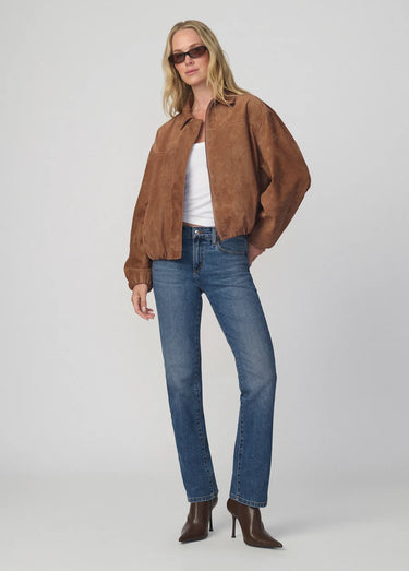 A woman modeling a brown suede-like cropped jacket over a white top, paired with blue jeans and brown heeled ankle boots, against a light gray studio background.