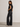 A woman wearing black wide-leg jeans, a black short-sleeve top, and white pointed-toe flats, standing in profile against a plain light gray studio wall.