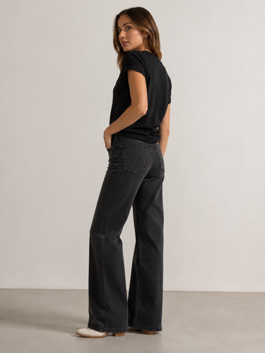 A woman wearing black wide-leg jeans, a black short-sleeve top, and white pointed-toe flats, standing in profile against a plain light gray studio wall.