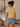Woman wearing a yellow t-shirt and denim shorts standing in a room with a plant and framed picture on the wall.