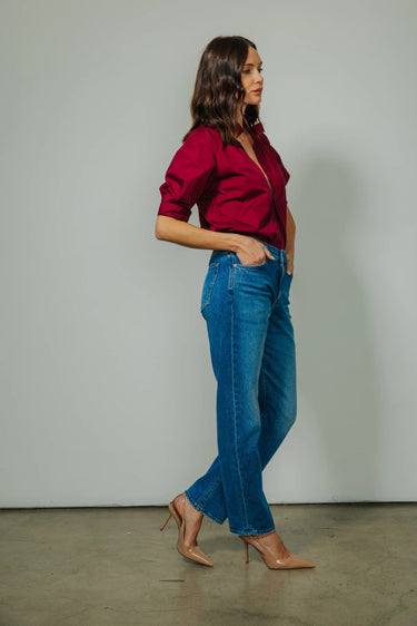A woman modeling a dark red button-down shirt and blue jeans with nude heels, standing in profile against a plain gray studio wall.