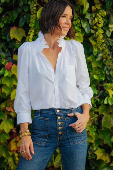 Woman wearing a white blouse and blue jeans standing against a green leafy background