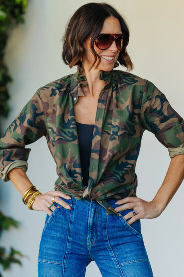 A woman wearing a vintage restructured men’s blouse in camo with white jeans on a neutral background.