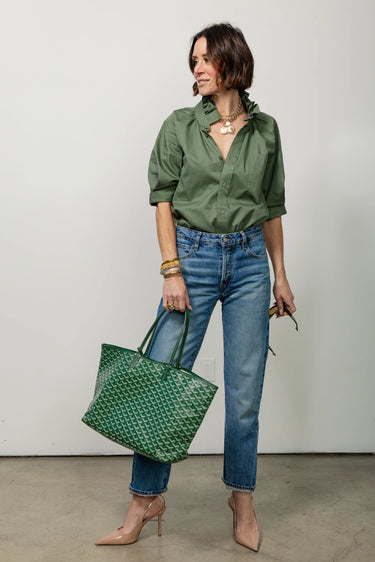 A woman wearing a restructured vintage men’s blouse in olive with a frill collar and blue jeans on a neutral background.