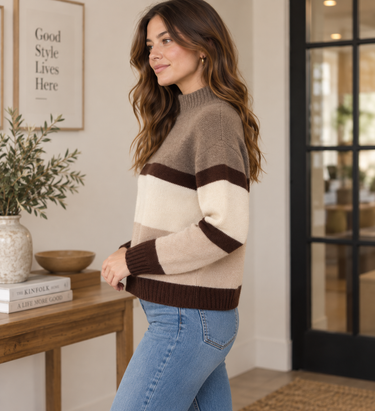 A woman wearing a color-block knit sweater with beige, cream, and dark brown horizontal stripes, paired with blue jeans, standing in a cozy, modern room.