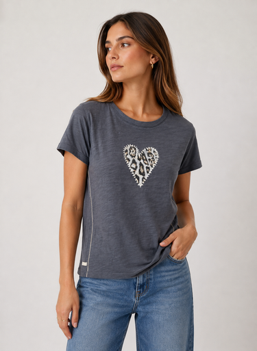 A woman wearing a dark gray heathered t-shirt with a white and black leopard-print heart graphic on the chest, paired with blue jeans, against a light gray studio background