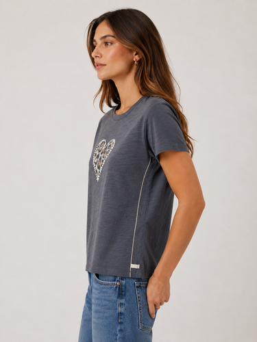 A woman wearing a dark gray short-sleeve t-shirt with a rhinestone-embellished heart on the chest, paired with blue jeans, shown from a side profile against a light gray studio background.