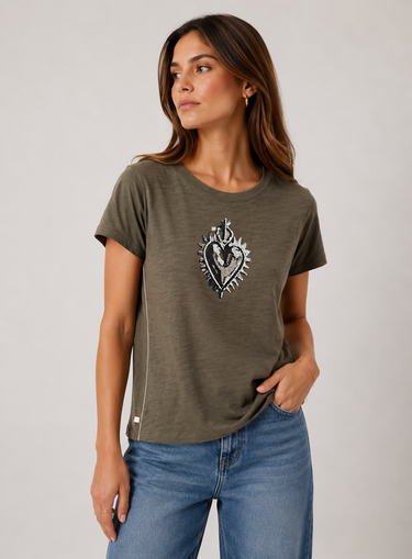 A woman wearing a short-sleeve crewneck t-shirt in olive green with a black and white graphic of a heart and cross on the chest, paired with blue jeans, photographed against a neutral light gray background.