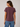 A woman shown from the back wearing a short-sleeve crewneck t-shirt in a heathered plum-brown color, paired with blue jeans, against a neutral light gray background.