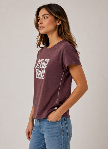 A woman wearing a short-sleeve crewneck t-shirt in a washed dark plum purple with a white graphic print on the front, paired with blue jeans, photographed against a neutral light gray background.