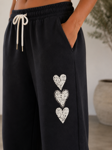Close-up of a person wearing black sweatpants with a white drawstring and three vertically stacked white heart motifs on the left leg, shown in a softly lit indoor setting.