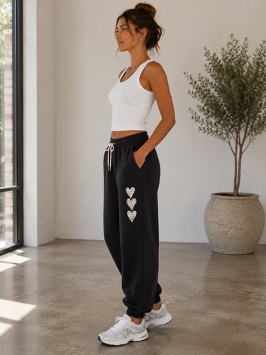 A woman stands in a bright indoor room wearing black jogger sweatpants with three silver heart embellishments down the side, paired with a white tank top and white sneakers.