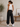A woman stands with her back to the camera wearing black wide-leg sweatpants with a small gold star patch on the pocket, a white ribbed tank top, and white sneakers in a bright indoor room.