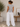 A woman shown from the back wearing a heather gray short-sleeve t-shirt and white wide-leg pants, standing in a minimalist room with a potted plant and a wooden side table.