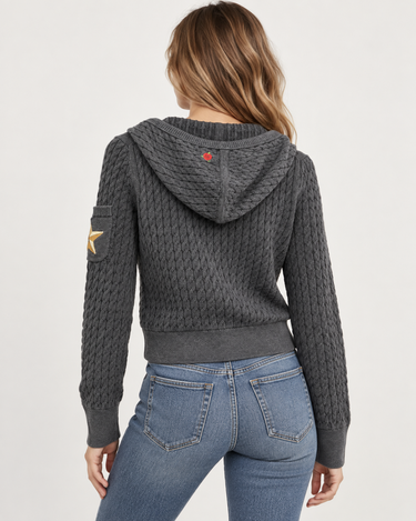A woman shown from the back wearing a dark gray cable-knit cropped hoodie with a hood, ribbed cuffs and hem, a small red logo patch on the hood, and a gold star patch on the sleeve, paired with blue jeans against a light gray studio background.
