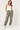 A woman modeling olive green jogger sweatpants with white embroidered stars down one leg, styled with a white tank top and white sneakers against a light gray studio background.