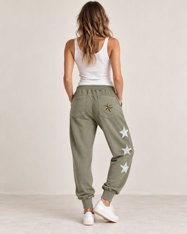 A woman viewed from behind wearing olive green jogger sweatpants with embroidered white stars down the side and a brown star patch on the back pocket, paired with a white tank top and white sneakers against a light beige studio background.