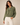 A woman wearing an olive green cropped utility jacket with a front zipper and flap chest pockets, paired with white pants, against a plain beige studio background.