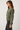 A woman shown in side profile wearing an olive green cropped utility jacket with front flap pockets and dark gray jeans, photographed against a plain light gray studio background.
