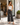 A woman stands on a sunlit sidewalk outside a clothing store, wearing dark charcoal wide-leg cropped pants with a white ribbed tank top and flat brown sandals.
