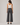 A woman viewed from the back wearing dark charcoal gray wide-leg cropped pants, a white ribbed tank top, and black flats, standing against a light gray studio background.