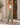 A woman stands in profile wearing olive green wide-leg pants, a white tank top, and beige block-heel mules on a sunlit porch with a potted plant and wooden door in the background.