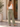 A woman stands with her back to the camera wearing high-waisted, wide-leg sage green corduroy pants, a white tank top, and brown block-heel mules in an outdoor patio setting.