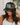 A woman wearing a camouflage trucker hat with a rectangular front patch that reads 'LAKE LIFE' in a blue and green graphic design. She is smiling and posing indoors with a white wall and green plant leaves in the background.