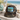 A camouflage trucker hat with a front patch reading 'LAKE LIFE' in green letters on a blue and white background, styled on a wooden surface with beach-themed props and a blurred ocean backdrop.