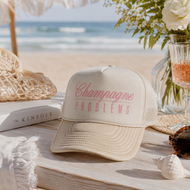 A cream-colored trucker hat with pink embroidered text reading 'Champagne PROBLEMS' sits on a wooden table in a beach-themed setting with a straw hat, sunglasses, a book, flowers, and the ocean in the background.