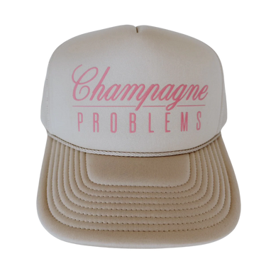 A beige trucker-style cap with a cream front panel and pink text reading 'Champagne PROBLEMS' on a white background.