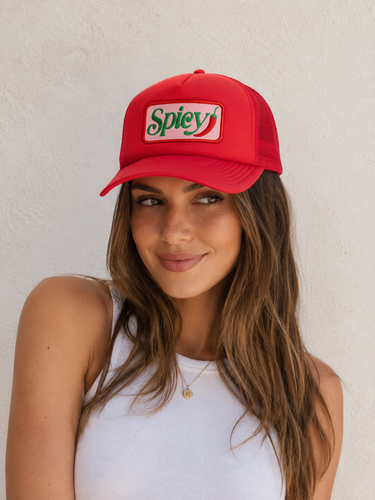 A woman wearing a red trucker-style cap with a white front patch that reads 'Spicy' in green script with a red chili pepper graphic, posed against a light textured wall.