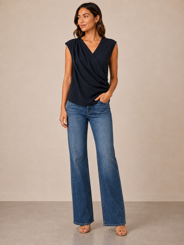 A woman modeling a sleeveless navy blue draped wrap-style top with blue flare jeans, standing against a plain beige studio backdrop.