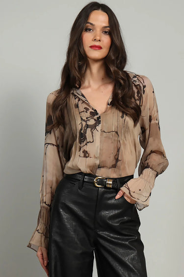 A woman wearing a v-neck chiffon printed brown blouse and black pants on a white background.