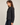 A woman wearing a dark washed denim jacket with a frayed hem and dark blue jeans, shown in a side profile against a plain light gray studio background.