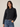 A woman modeling a black cropped denim jacket with a button-down front, chest flap pockets, and a ruffled hem, paired with blue jeans against a plain light beige studio background.