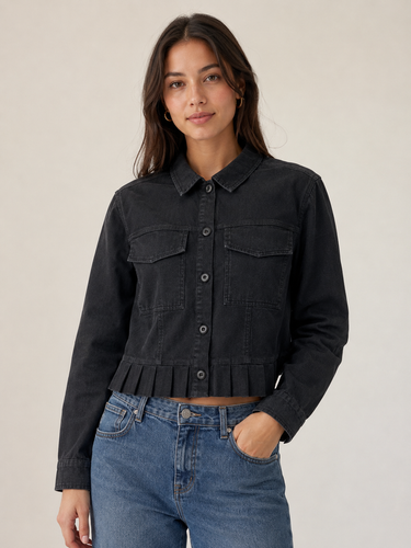 A woman modeling a black cropped denim jacket with a button-down front, chest flap pockets, and a ruffled hem, paired with blue jeans against a plain light beige studio background.